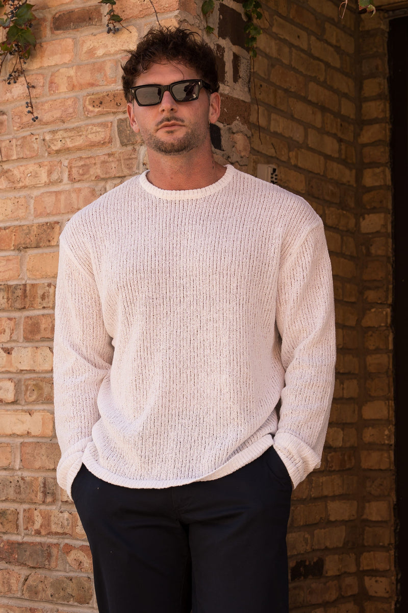 A man wearing a cream-colored ribbed knit sweater and dark sunglasses, standing against a warm brick wall corner.