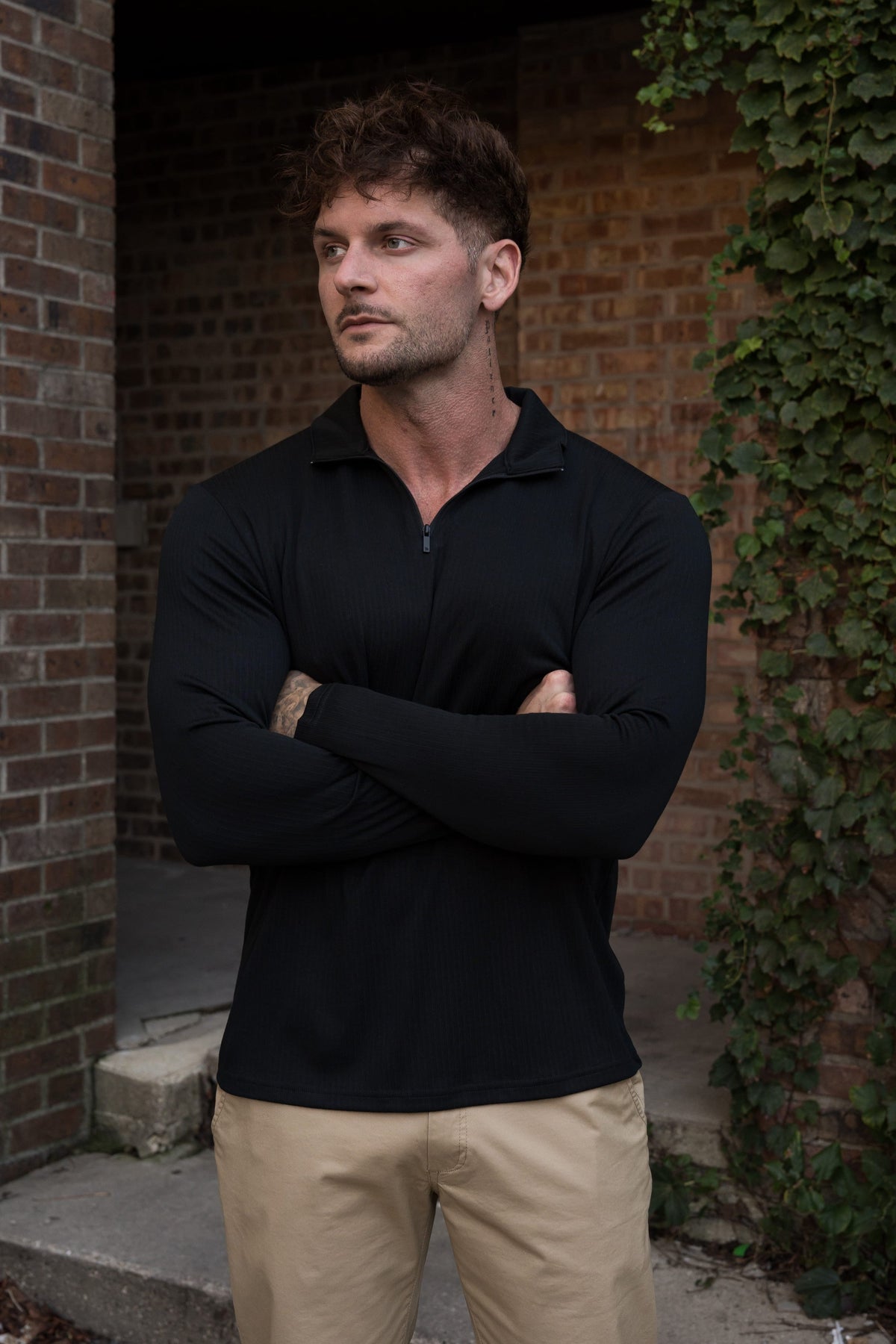 A man wearing a black long-sleeve quarter-zip polo shirt and khaki pants, standing with arms crossed in front of a brick wall with ivy.