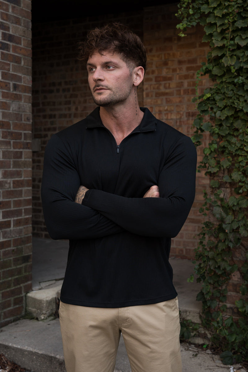 A man wearing a black long-sleeve quarter-zip polo shirt and khaki pants, standing with arms crossed in front of a brick wall with ivy.